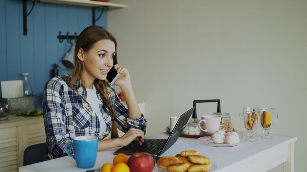Freelancer working from her kitchen on a laptop while taking a phone call.