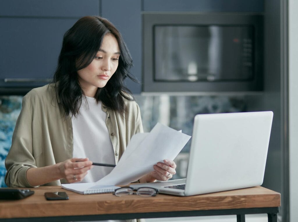 Person at a kitchen table at night reviewing a pay stub and a tax form by laptop light